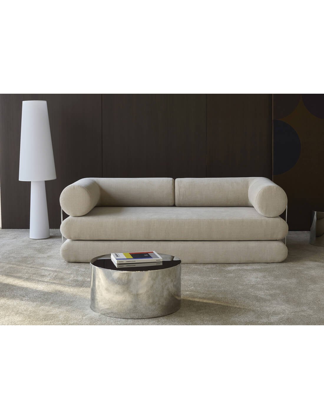 Canapé beige moderne à épaules cylindriques avec ottomans, devant mur sombre et table basse en métal brillant. Canapé beige moderne à épaules cylindriques avec ottomans, devant mur sombre et table basse en métal brillant.