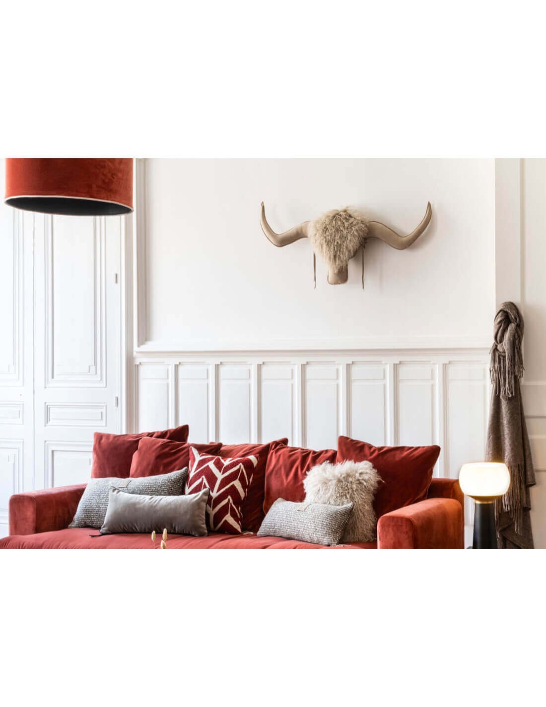 Canapé rouge élégant avec coussins gris et à motifs, mur blanc mouluré et décoration tête de bœuf sur le mur, ambiance déco chaleureuse.