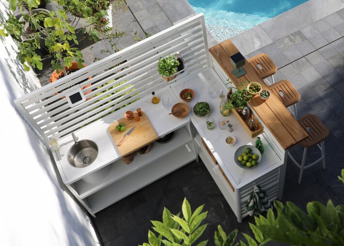 Cuisine extérieure en L avec îlot et plan de travail compact, préparations et fruits, mobilier en bois et plantes, raccordée à une piscine. Aménagement cuisine petit espace optimisé.